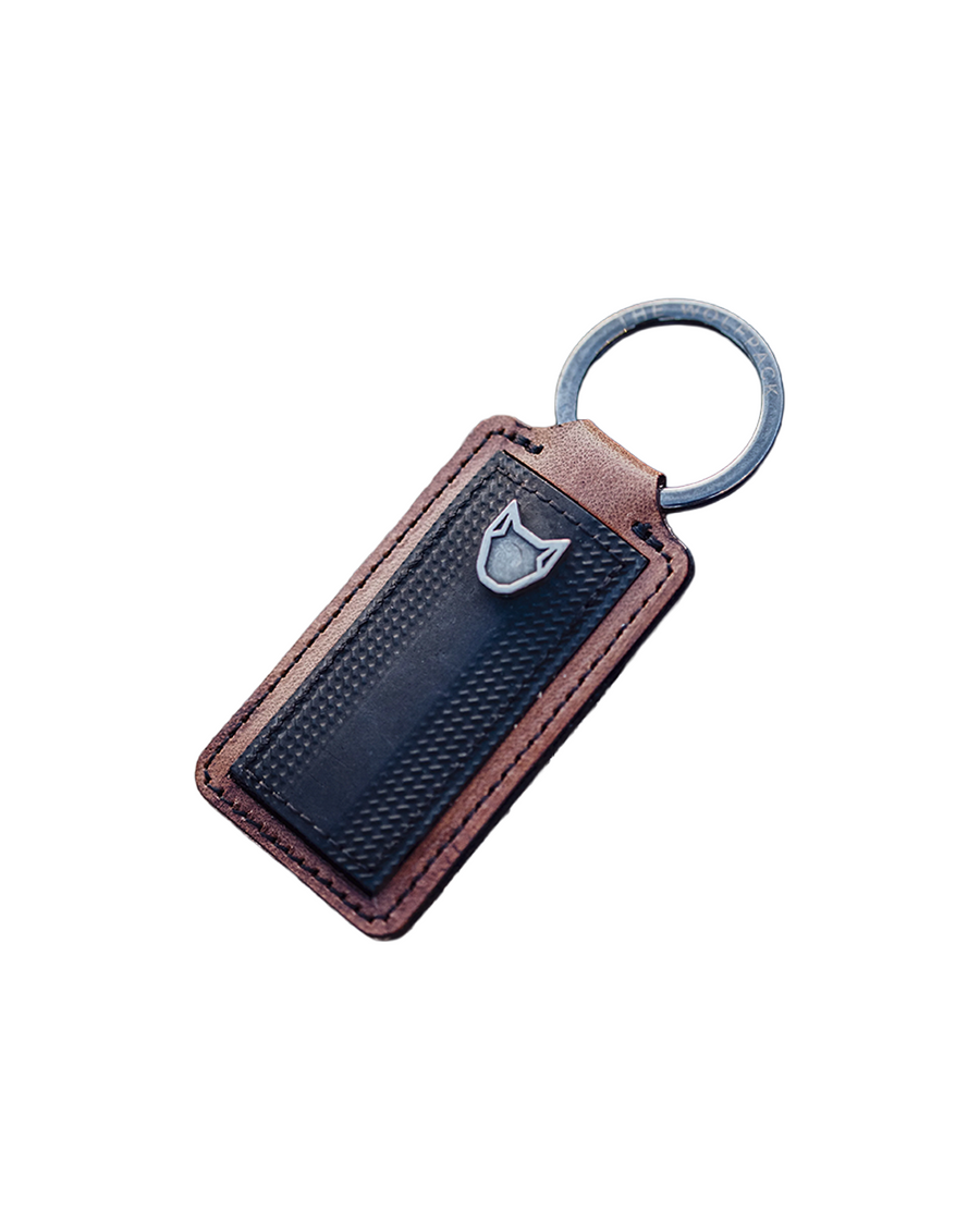 A rectangular leather keychain with a dark textured center panel, brown leather edges, a small silver shield emblem, and a silver key ring, shown on a plain white background.