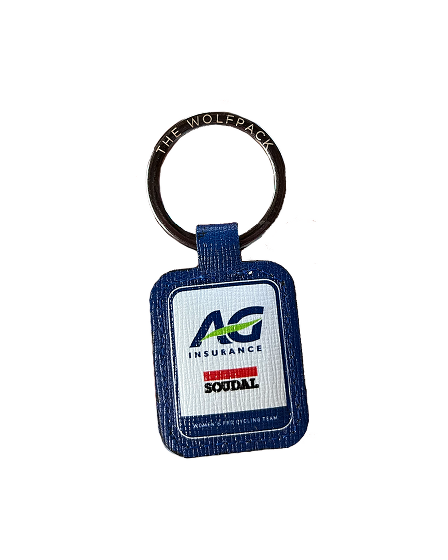 A blue textured leather keychain featuring a white rectangular patch with the 'AG Insurance' and 'Soudal' logos, attached to a metal ring that reads 'THE WOLFPACK'.