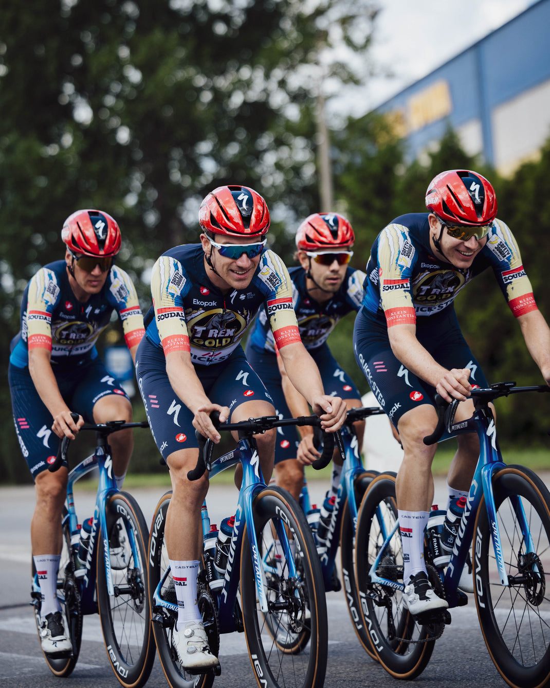 Four cyclists riding in a peloton on a road bike team, wearing matching navy, cream, and red T-rex Quickstep jerseys with red Specialized helmets and sunglasses, captured outdoors on a road with trees and a building in the background.